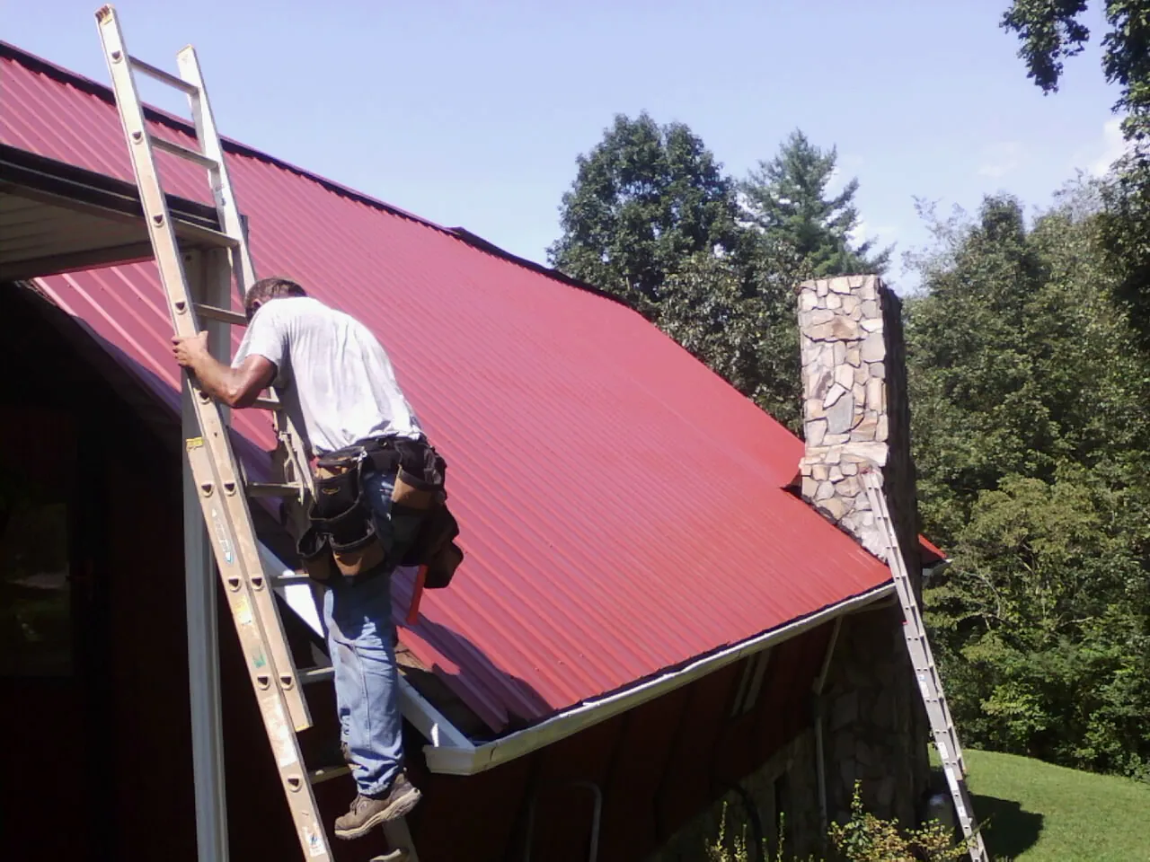 Roofer on a ladder beside a red metal roof and stone chimney — Soffit & Fascia Repair by Roofing Brothers in Salley